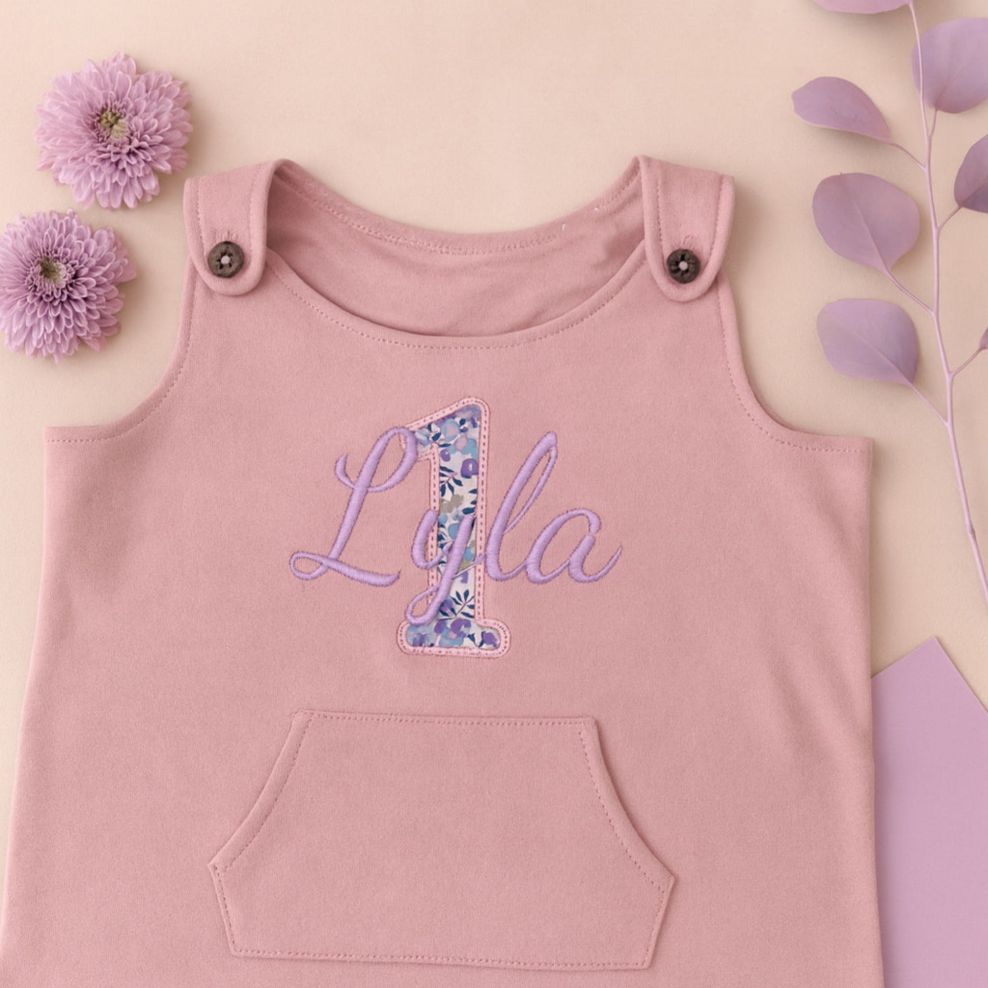 Pink baby romper with 'Lyla 1' on a light pink background with flowers and leaves.