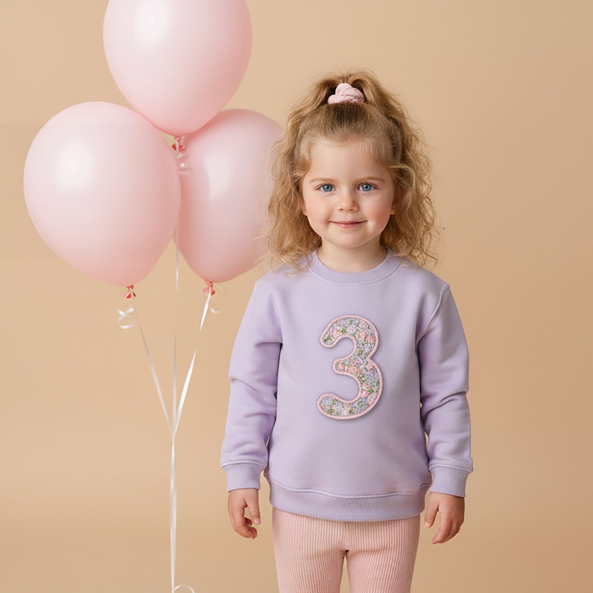 Child wearing a purple sweatshirt with the number 3, holding pink balloons against a beige background