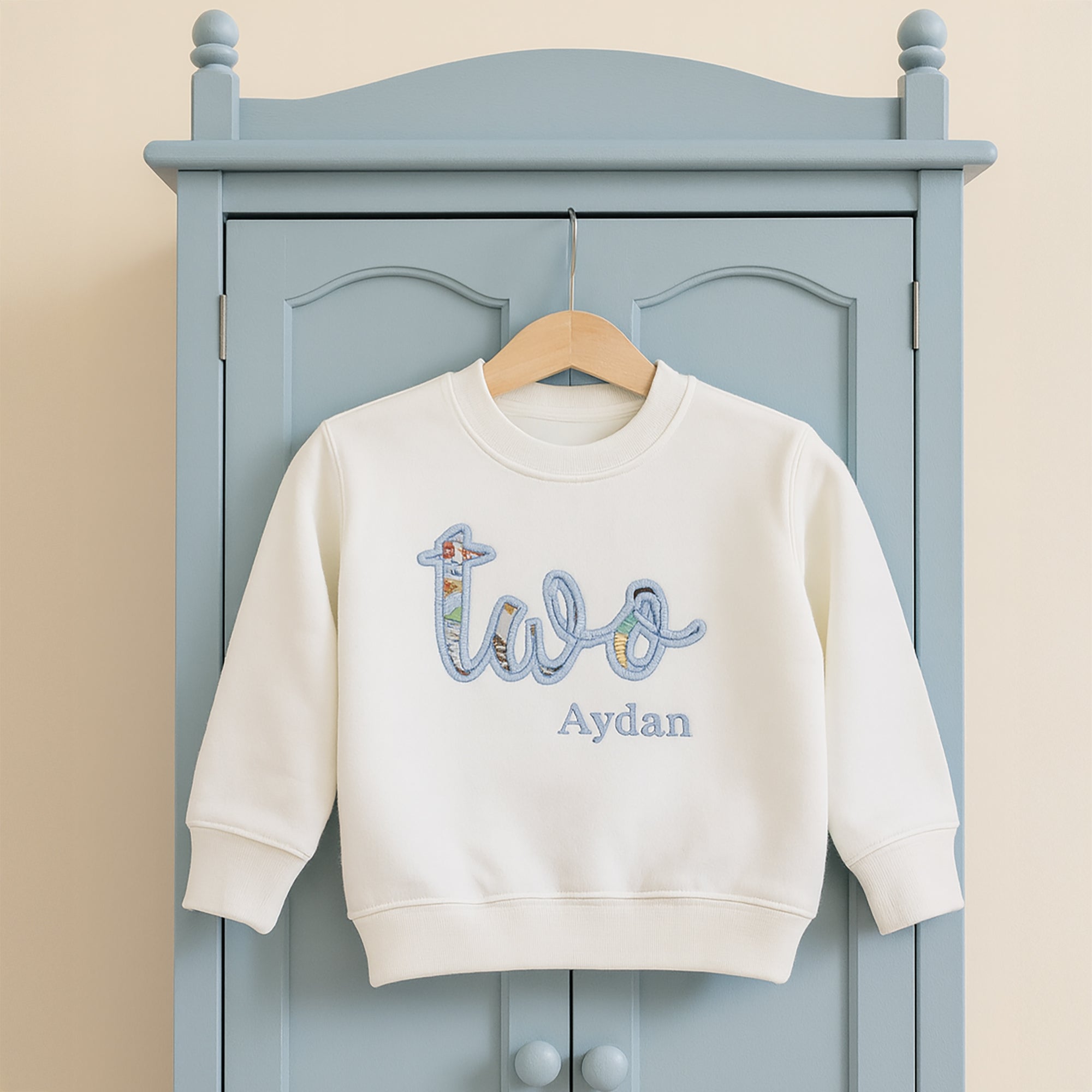 White sweatshirt with 'two Aydan' text on a hanger against a blue cabinet.