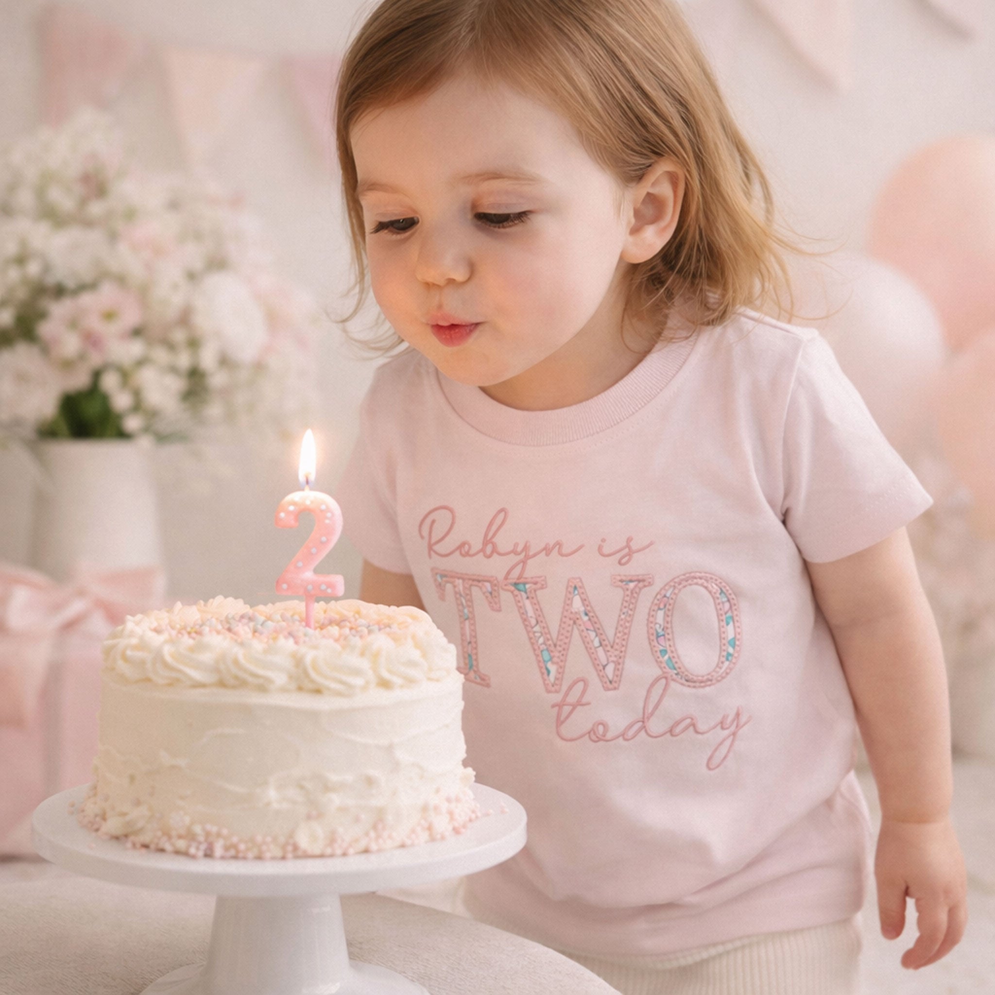 Child blowing out a candle on a birthday cake with 'Rabyn is TWO today' shirt
