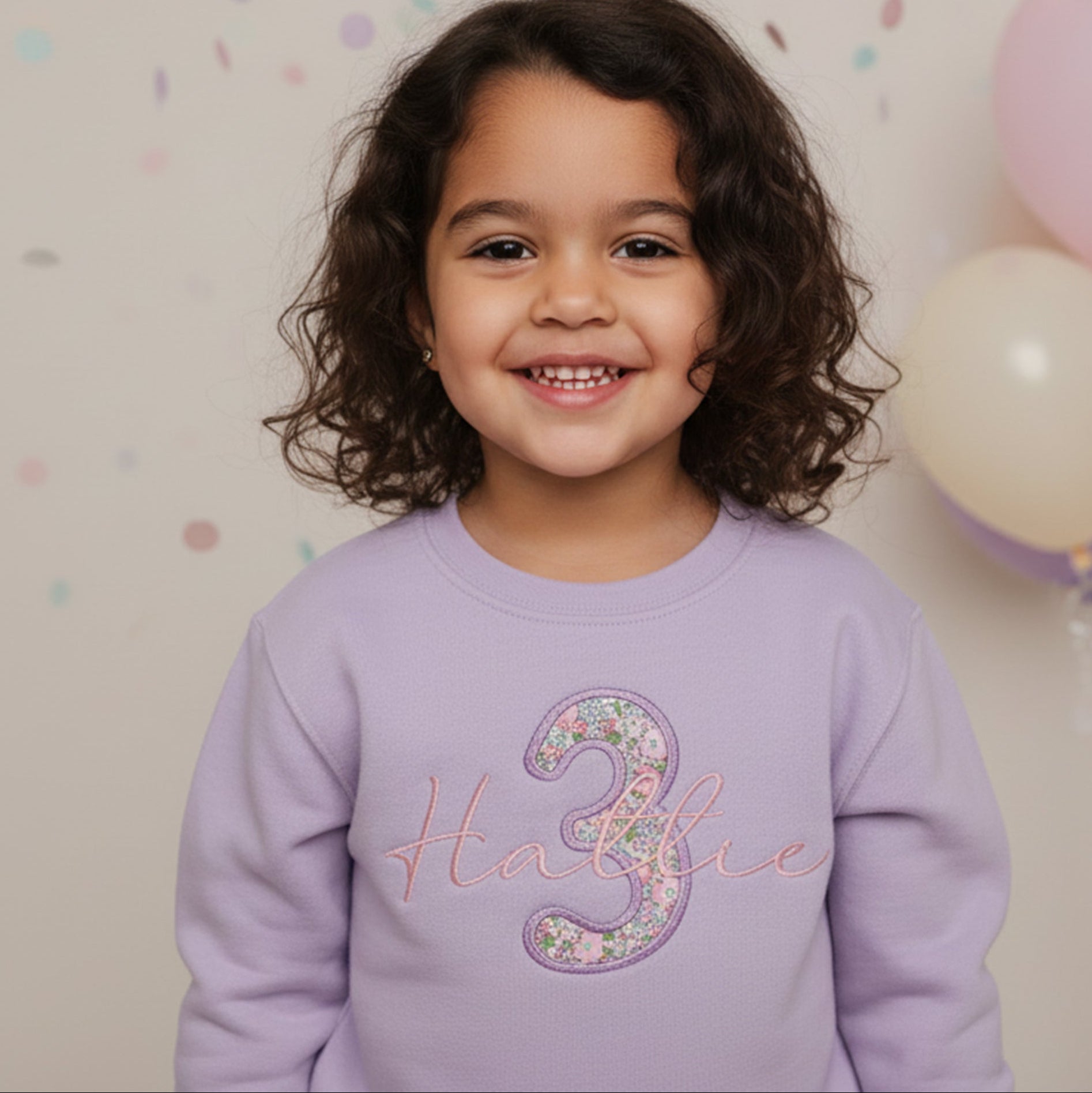 Child wearing a purple sweatshirt with 'Hattie 3' on it, standing in front of balloons.