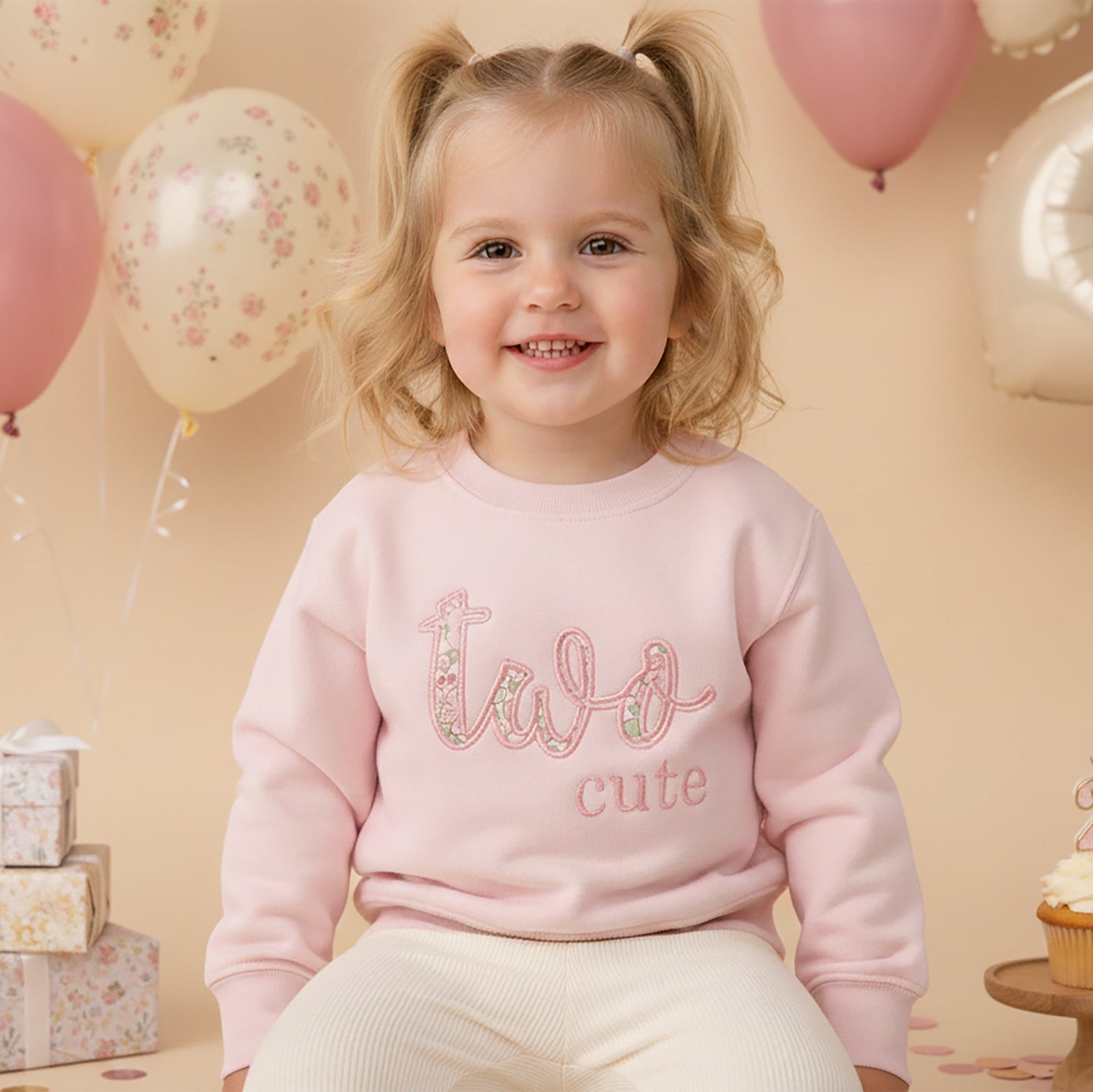Child wearing a pink sweatshirt with 'two cute' text, surrounded by balloons and gifts on a beige background.