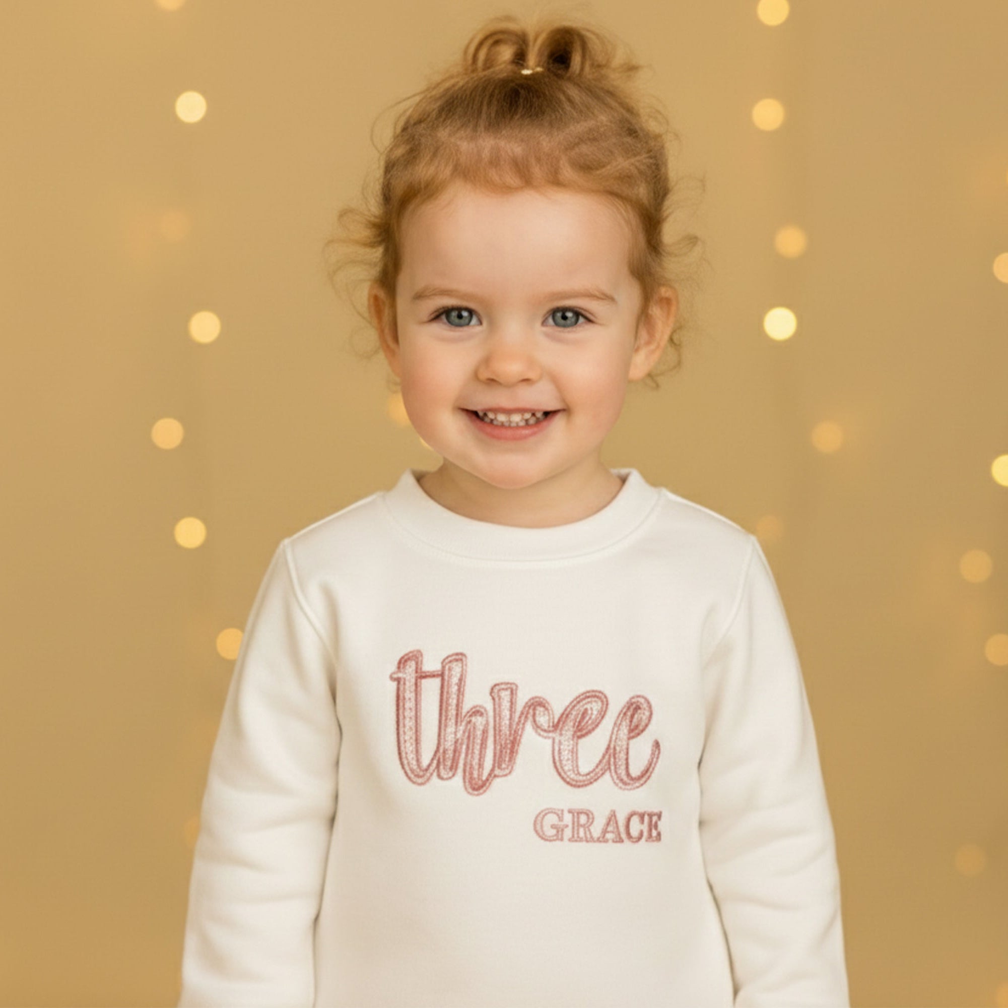 Child wearing a white personalised  third birthday sweatshirt with 'Three Grace' text against a gold background with lights
