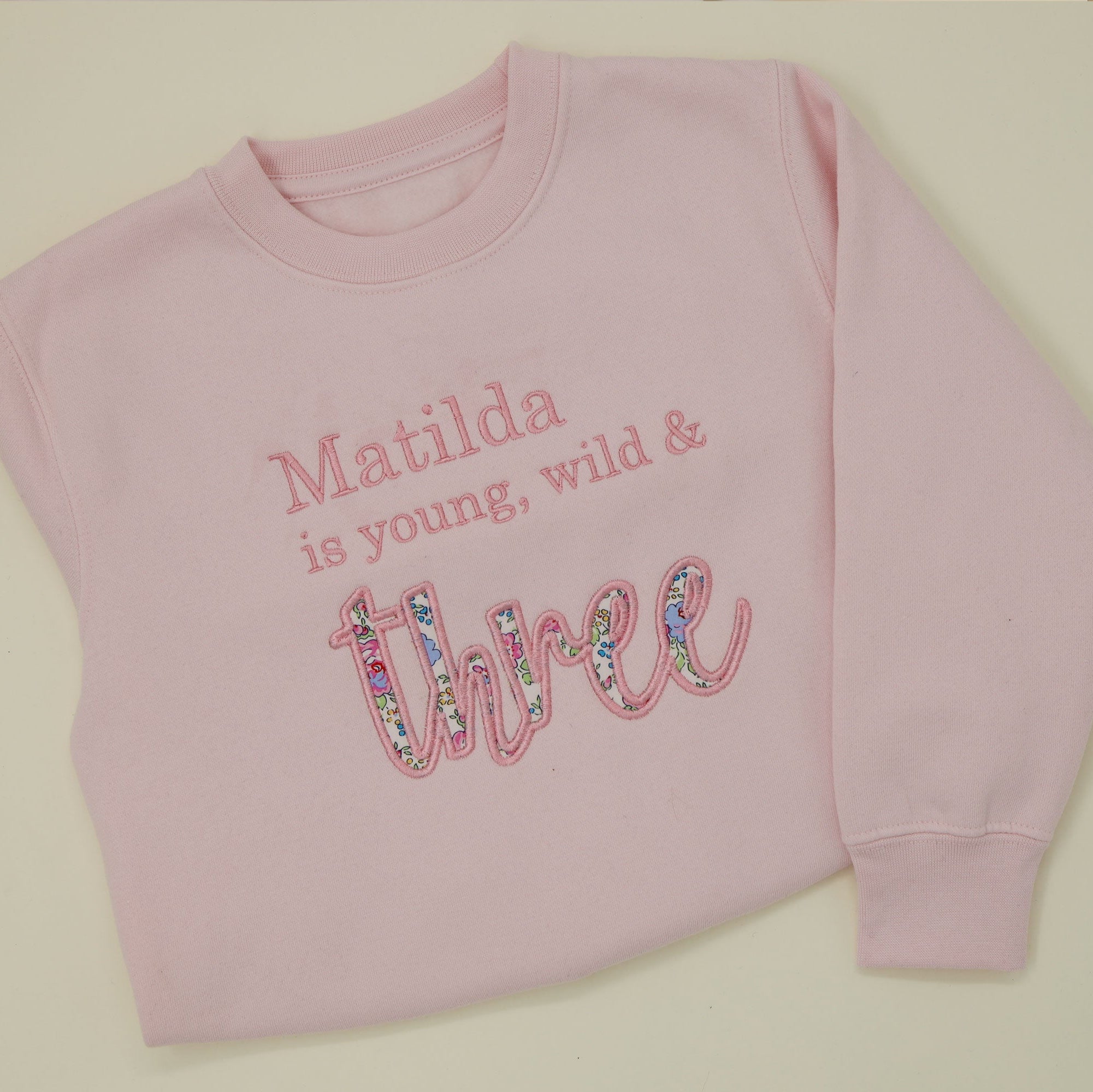 Pink personalised 3rd birthday jumper with text 'Matilda is young, wild & three' on a light background